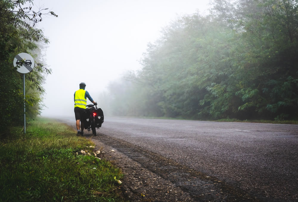 Cyclist with reflective vest and lighs on is walking down the road to the fog in nature. Bicycle touring in bad weather conditions and bad visability