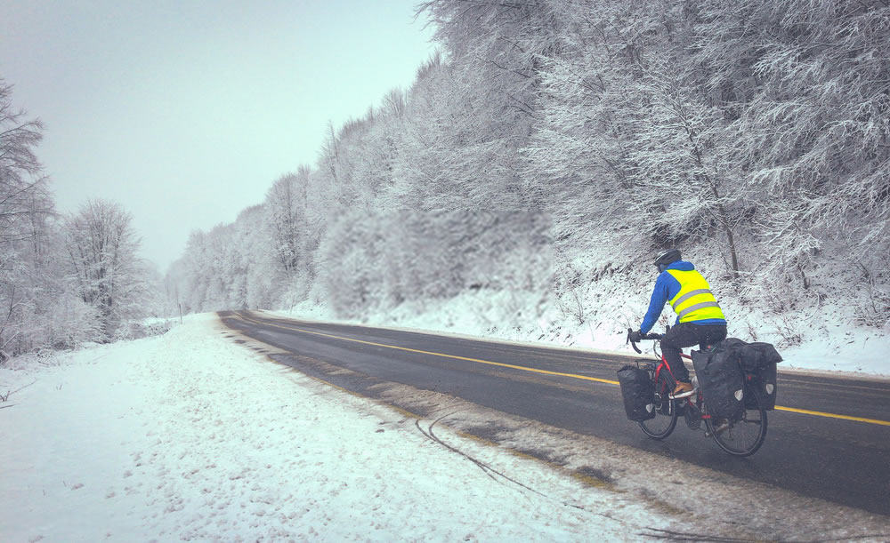 Male man in black and bue is cycling on fully loaded touring bicycle in winter conditions in nature covered in snow on wet asphalt with no cars